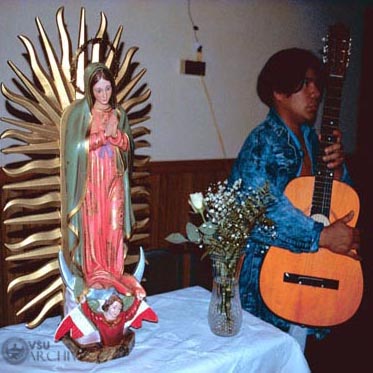 Latino guitarist next to a Virgin Mary statue.