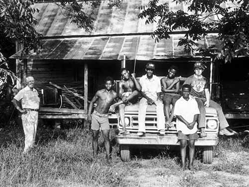A tobacco crew sits on an old truck outside of an old house.