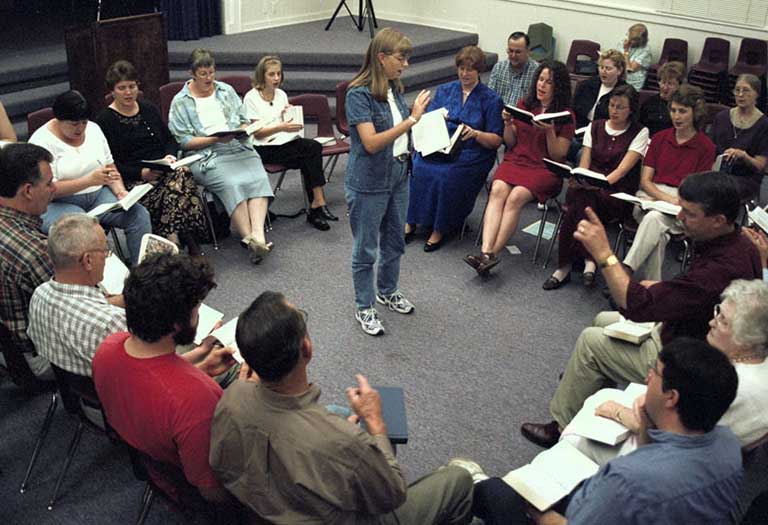 A woman stands leading a sing from the middle of a seated group.