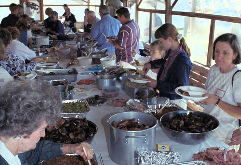 A crowd makes their plates of food. A large feast is laid out on a long table.