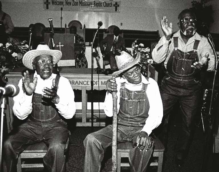 Black and white photograph of old black men in overalls siting or standing, while clapping and singing in a church.